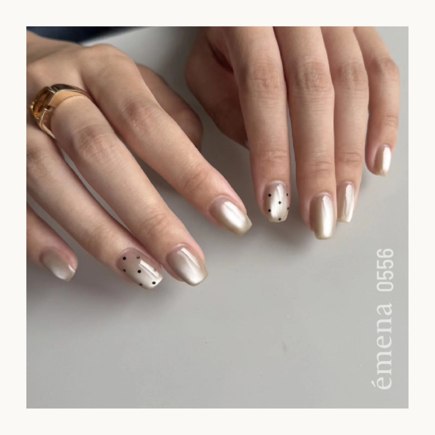 Close-up of hands with beige nail polish and a ring on a gray background, featuring the brand 'emena'.