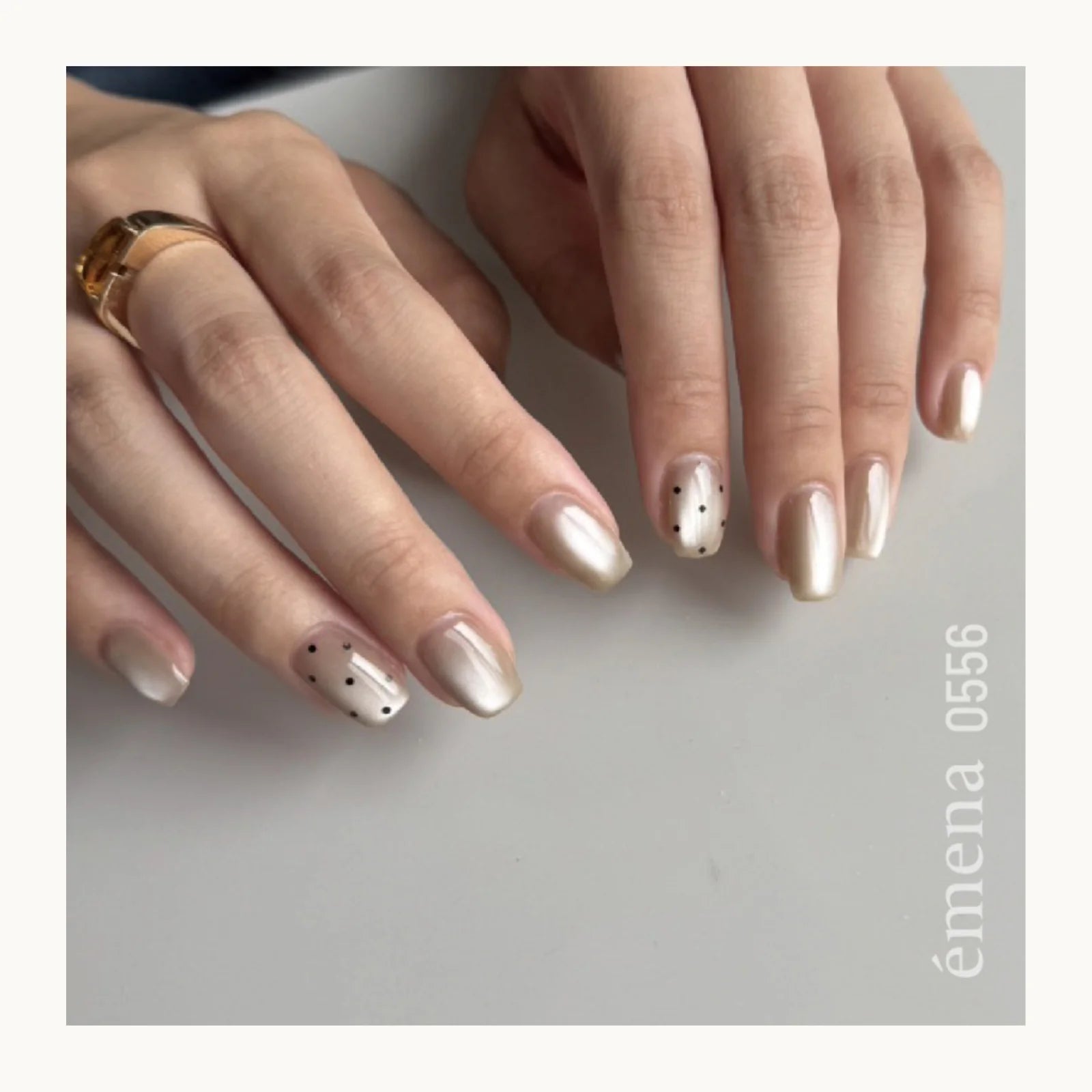 Close-up of hands with beige nail polish and a ring on a gray background, featuring the brand 'emena'.